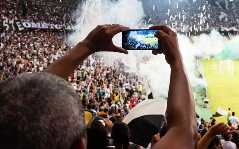 Fan in a packed stadium holding up a smartphone to film the pitch as smoke flares drift over the cheering crowd