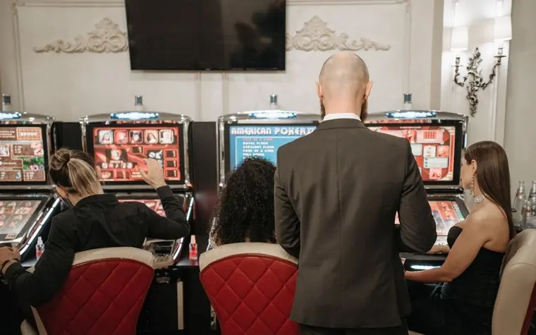 Casino patrons seated at upright video gaming machines with a pit attendant standing behind them