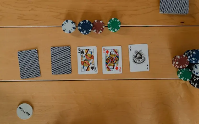 Two queens and an ace dealt face up on a felt table beside stacked casino chips and a dealer button