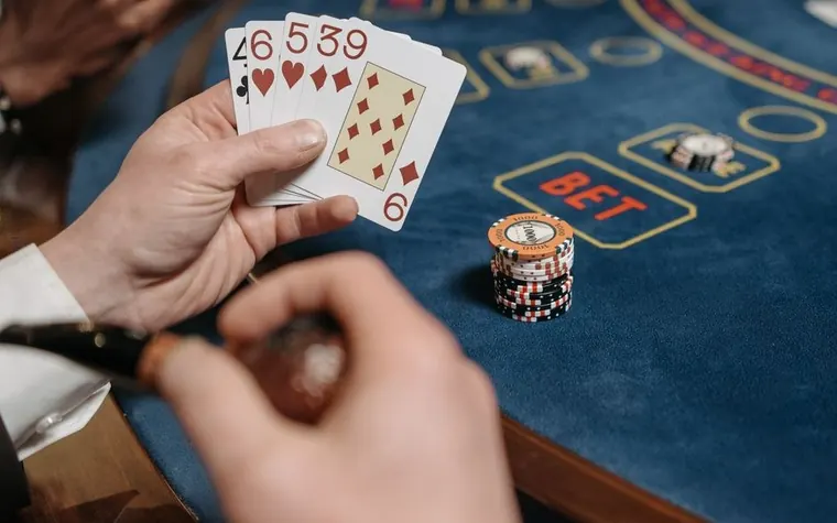 A player's hand holding five playing cards, a four, two sixes, a five, and a nine, beside a stack of casino chips on a blue felt betting table