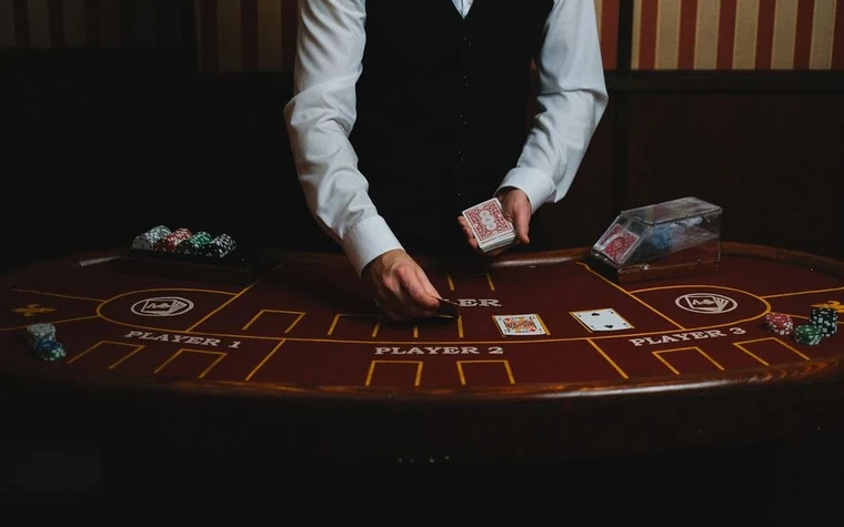 Dealer in a white shirt and black vest dealing cards across a dark curved casino table with printed betting areas