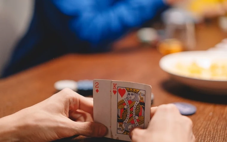 a hand holding a king of hearts and another playing card over a wooden table with chips blurred in the background
