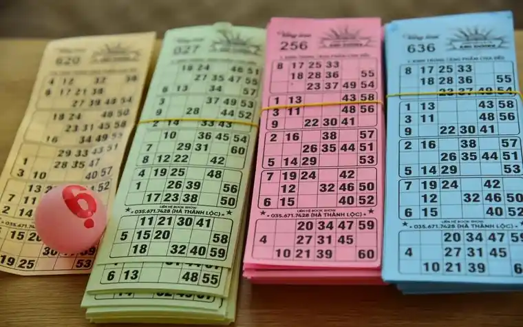 four colored paper bingo cards in yellow, green, pink and blue with printed number grids lined up on a wooden table, with a pink numbered ball beside them