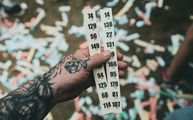 hand with a forearm tattoo holding two paper strips printed with columns of numbers over a blurred background of scattered colorful confetti