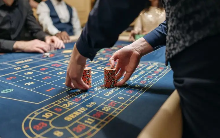 two hands moving stacks of red casino chips across the blue betting layout of a roulette table with a dealer in a vest standing nearby