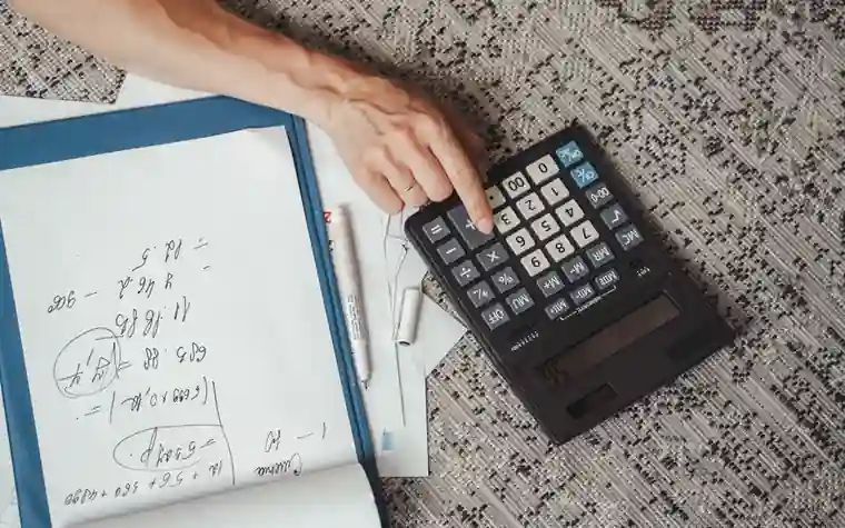 overhead view of a hand pressing keys on a black desktop calculator beside a notepad covered in handwritten calculations
