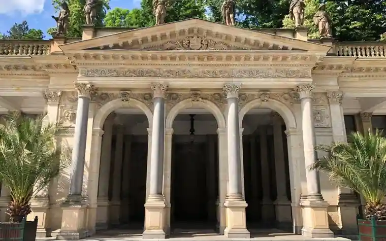 Stone colonnade with tall columns and arched openings beside palm plants at a historic spa pavilion