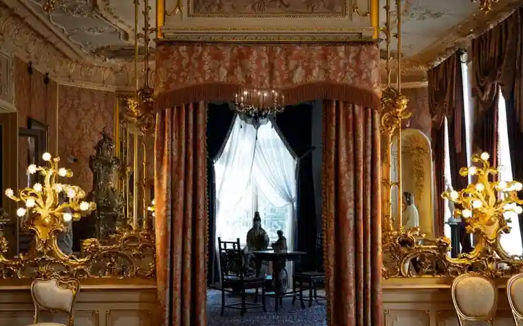 Gilded salon with draped curtains, gold candelabra, and a chandelier seen through an arched doorway
