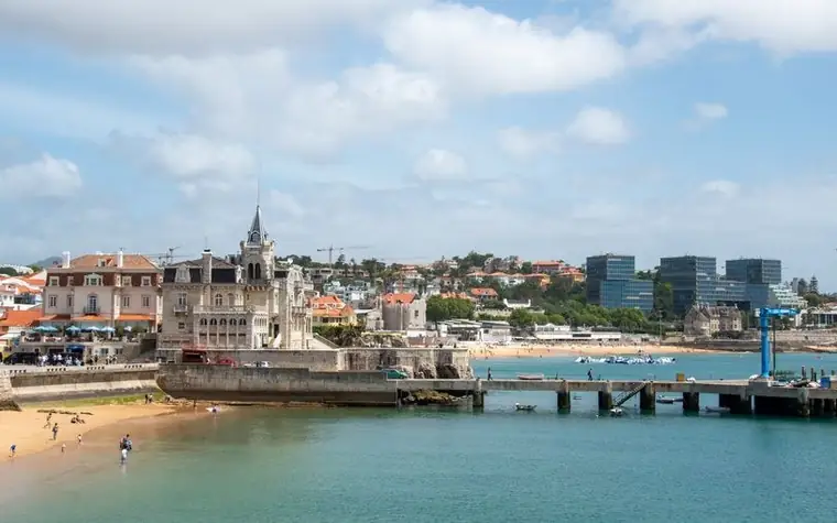 Cascais waterfront with sandy beach, small pier, and pastel seaside buildings under a blue sky