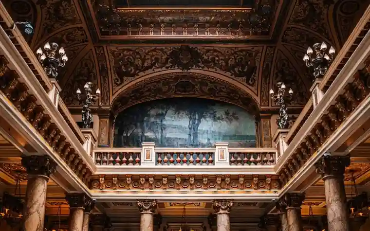 Ornate gilded hall with carved columns, a decorated balcony, and hanging lamps