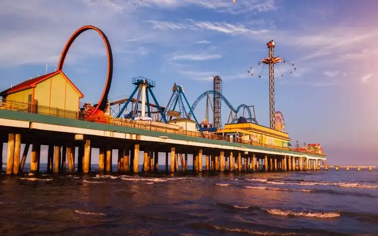 Seaside amusement pier at sunset with a looping roller coaster and tall rides rising over the water