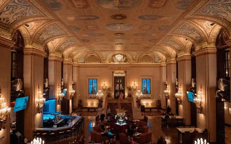 Ornate hotel lobby with a gilded coffered ceiling, hanging chandeliers, and a seating area below