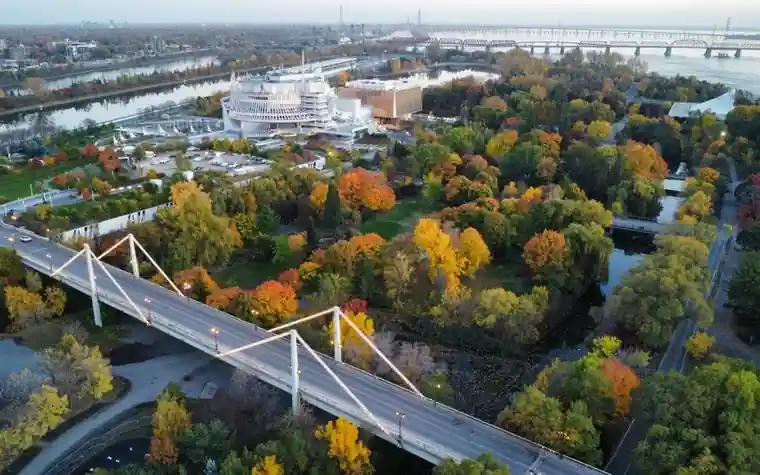Aerial view of an island park in autumn colors with a footbridge, a river channel, and large buildings among the trees