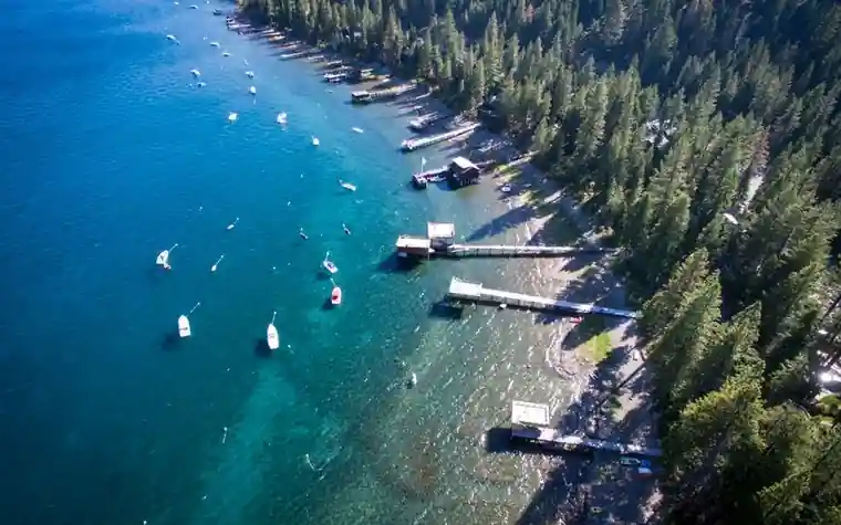 Aerial view of a forested lake shoreline with clear blue-green water, small boats, and wooden docks along the pine-covered coast