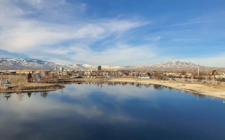 City skyline with a few mid-rise buildings reflected in a calm lake, with snow-dusted mountains and a pale blue sky behind it