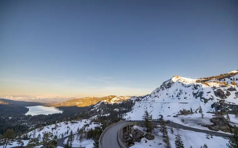 Snow-covered mountain pass at dusk with a winding road in the foreground and a distant lake in the valley below