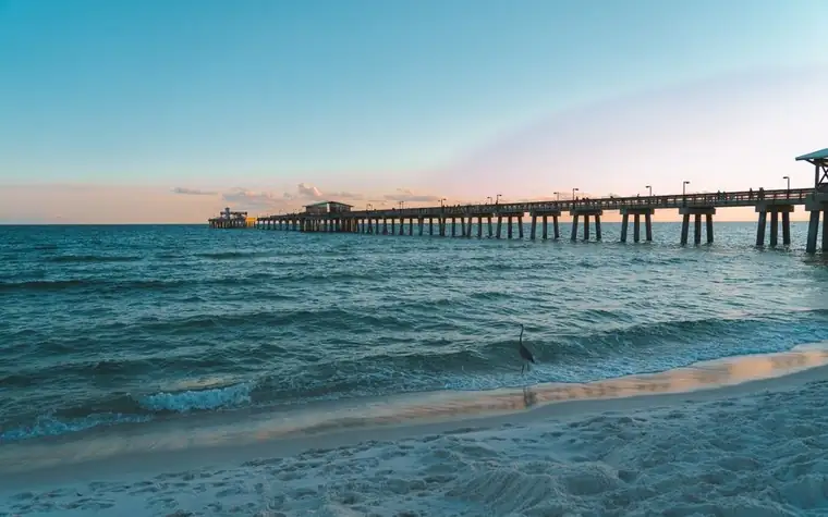 Long wooden pier stretching out over turquoise surf toward the horizon, with wet sand and foam breaking in the foreground