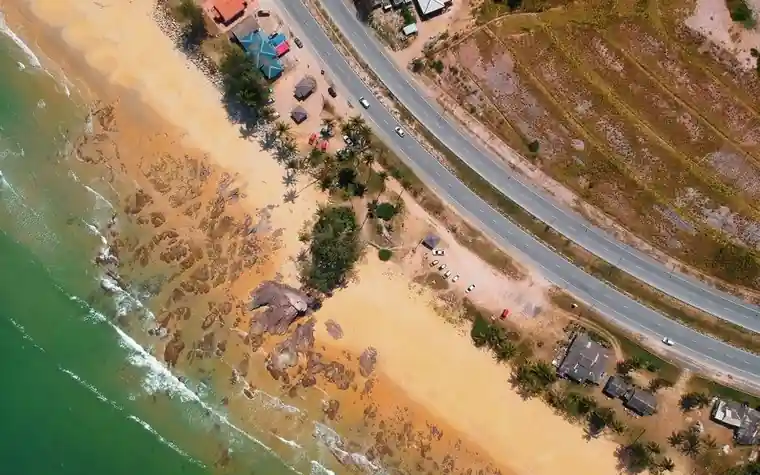 Aerial view of a coastal road running beside a sandy beach with green surf, scattered trees, and small buildings