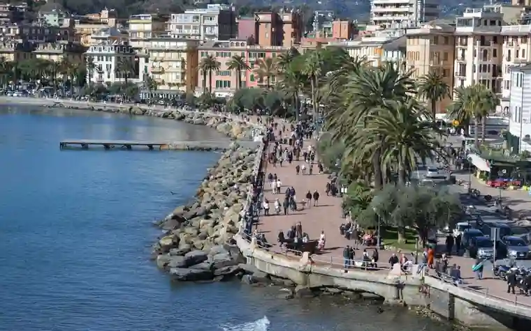 curved seaside promenade along a rocky breakwater with palm trees, pedestrians and apartment buildings climbing a hillside above the water