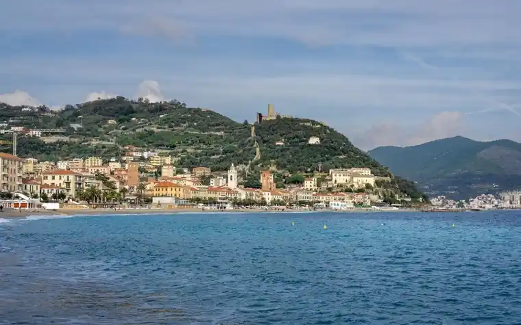 view across blue water to a hillside Ligurian coastal town with pale buildings, a hilltop tower and mountains rising in the distance