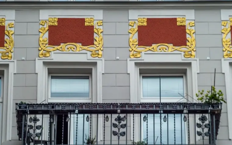 ornate Art Nouveau building facade in grey and white with gold floral plaster ornaments around red panels above a wrought-iron balcony