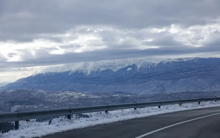 Snow-covered mountain ridge seen from a highway guardrail under a cloudy winter sky