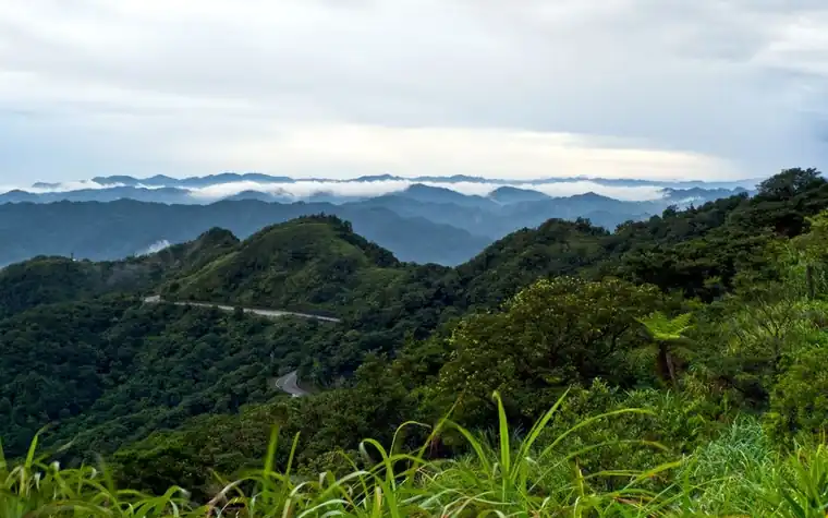 Winding two-lane mountain road cutting through green forested hills with clouds settled in the valleys