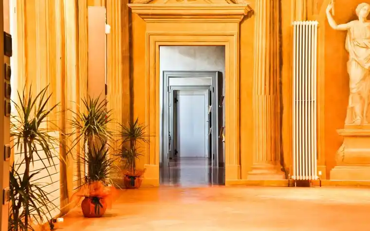 long ornate hallway with classical columns, potted plants, and a statue leading toward a doorway