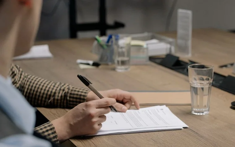 close-up of a hand signing a paper document with a pen beside a glass of water on a wooden desk