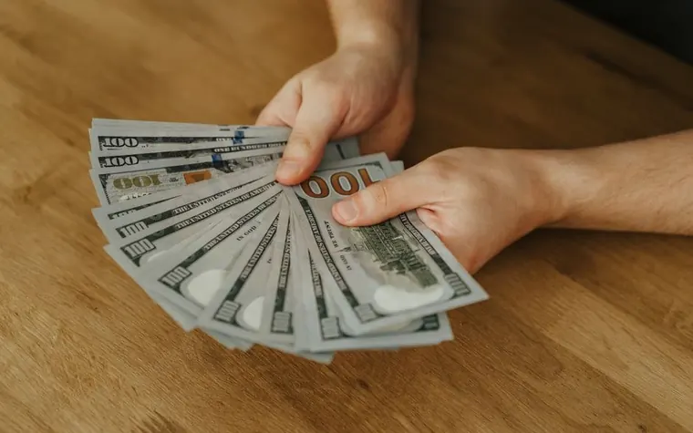 hand holding a fanned stack of hundred dollar bills over a wooden tabletop