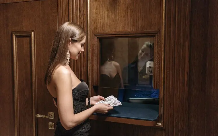 woman in a black dress standing at a wooden casino cashier window with a glass partition and a small counter tray