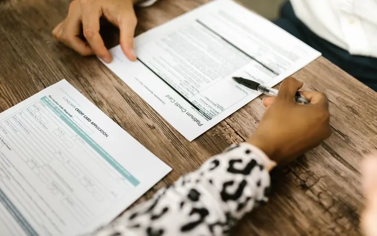 two people's hands filling out printed forms on a wooden table with pens