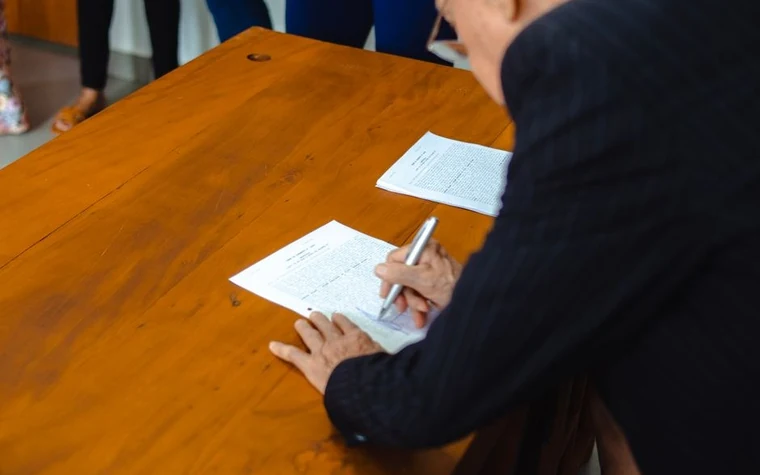person in a dark suit signing a paper form on an orange wooden table