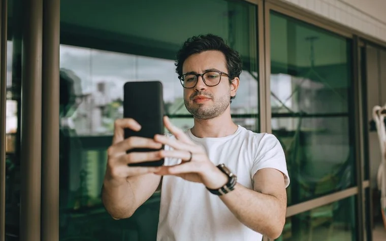 man in a white t-shirt and glasses holding up a black smartphone to take a photo near a glass window
