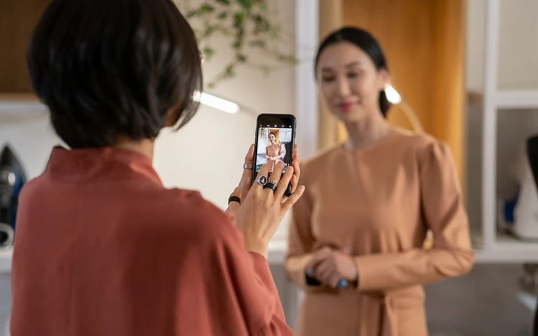 woman in an orange top holding a smartphone to photograph another woman in a bright room