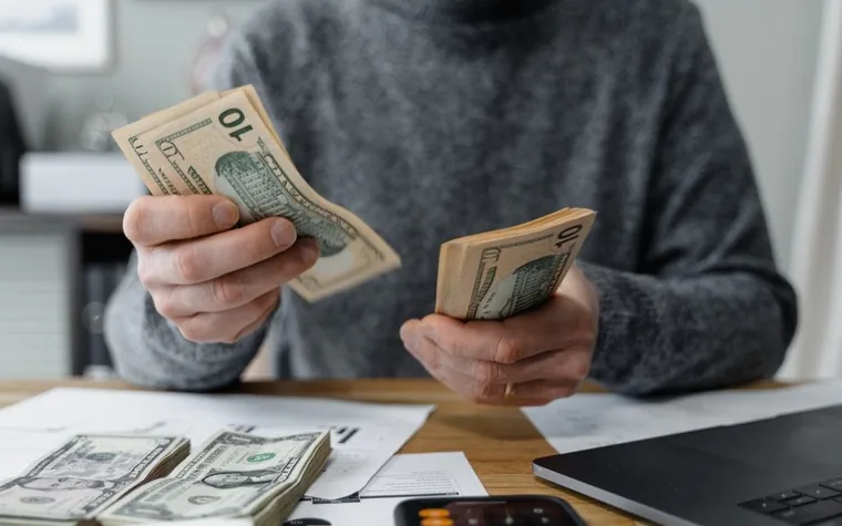 man in a gray sweater counting a stack of US dollar bills at a wooden desk