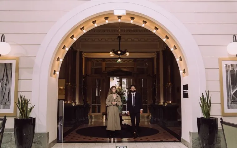 couple in long coats standing under a lit archway at the entrance of an elegant hotel lobby