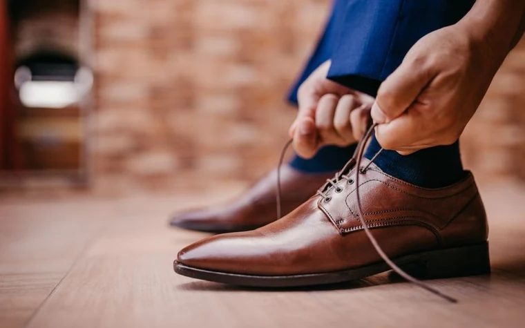hands tying the laces of polished brown leather dress shoes on a wooden floor