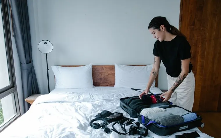 woman in a black top packing clothes into an open suitcase on a white hotel bed