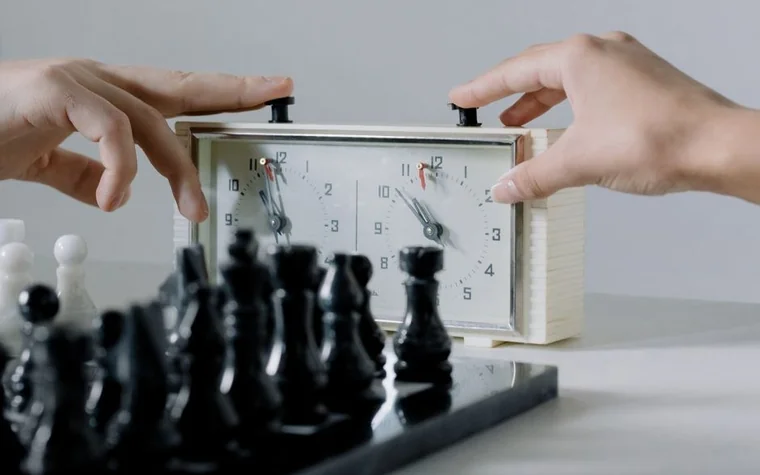 two hands pressing buttons on a chess clock above a chessboard with black pieces