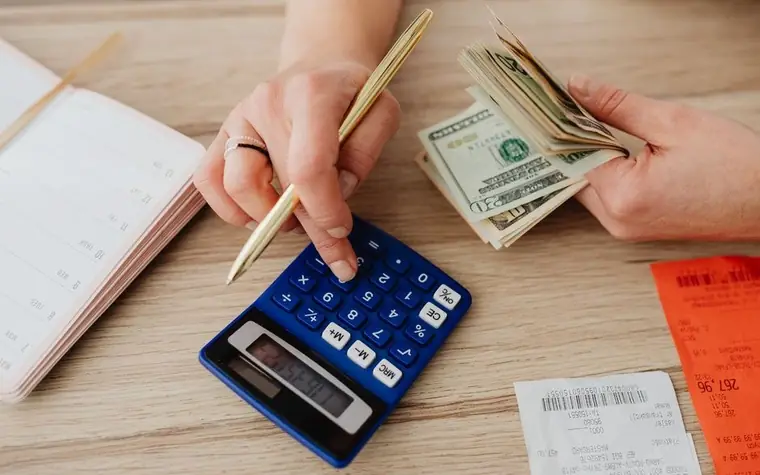 Hands holding a fold of US dollar bills over a blue calculator, notebook and receipts