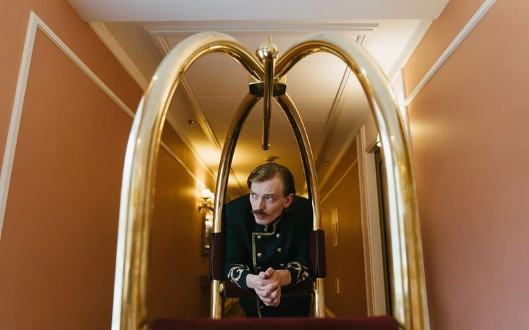 Bellhop in a vintage gold-trimmed uniform pushing a luggage cart through a hotel hallway