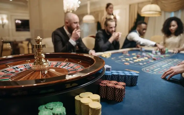 roulette wheel and stacked casino chips on a gaming table with several players seated around it