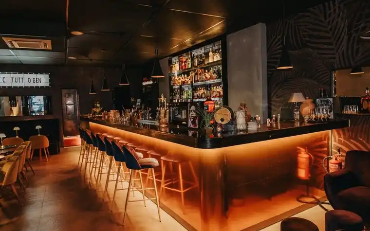 a dimly lit hotel bar with an illuminated amber counter, high stools along the front and rows of bottles on shelves behind it
