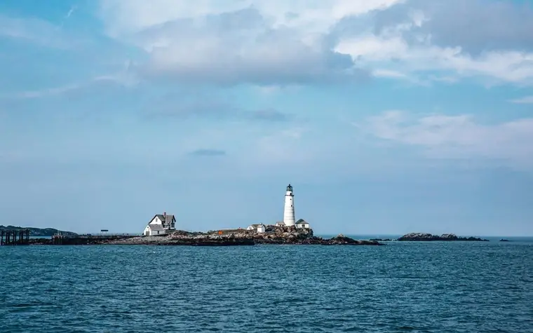 a white lighthouse on a small rocky island far out on calm blue water under a cloudy sky
