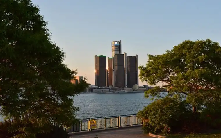 Detroit's Renaissance Center towers reflected across the river at dusk, framed by waterfront trees