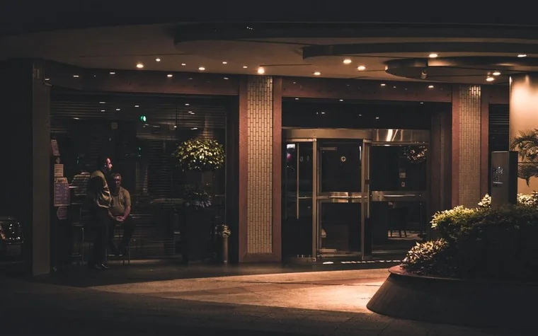 Hotel entrance at night with lit glass doors and two people standing near the doorway