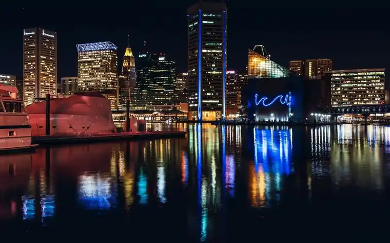 Baltimore skyline at night with lit office towers and boats reflected in the dark water of the harbor