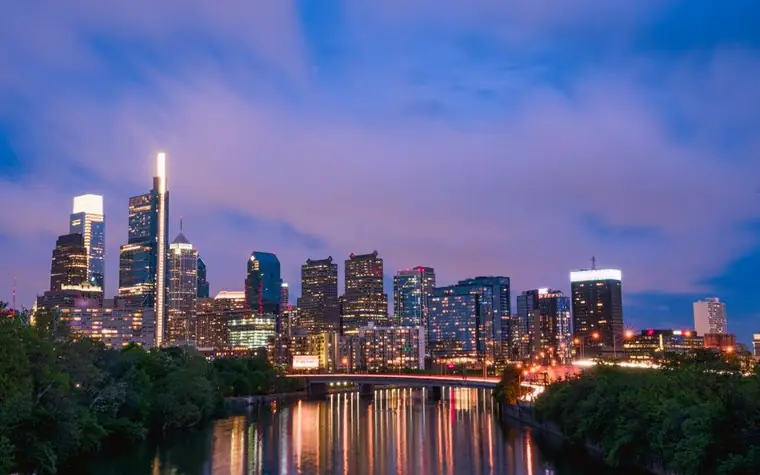 Philadelphia skyline with glass towers lit at dusk above a river and a bridge with the city's lights reflected in the water