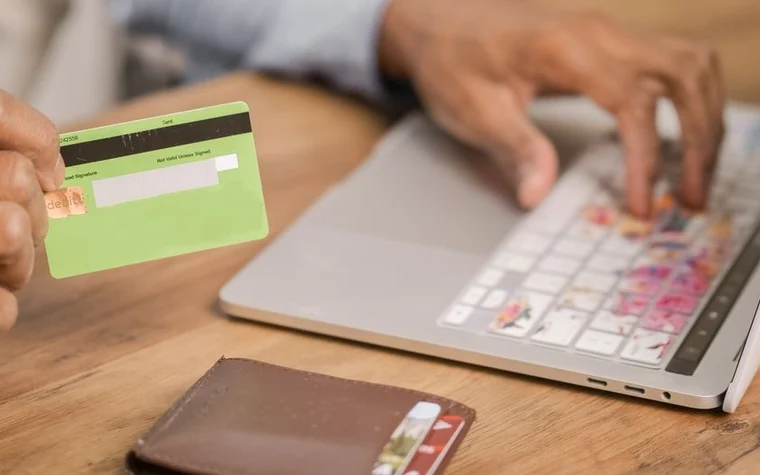 hand holding a green plastic card above a laptop keyboard next to a brown leather wallet on a wooden desk